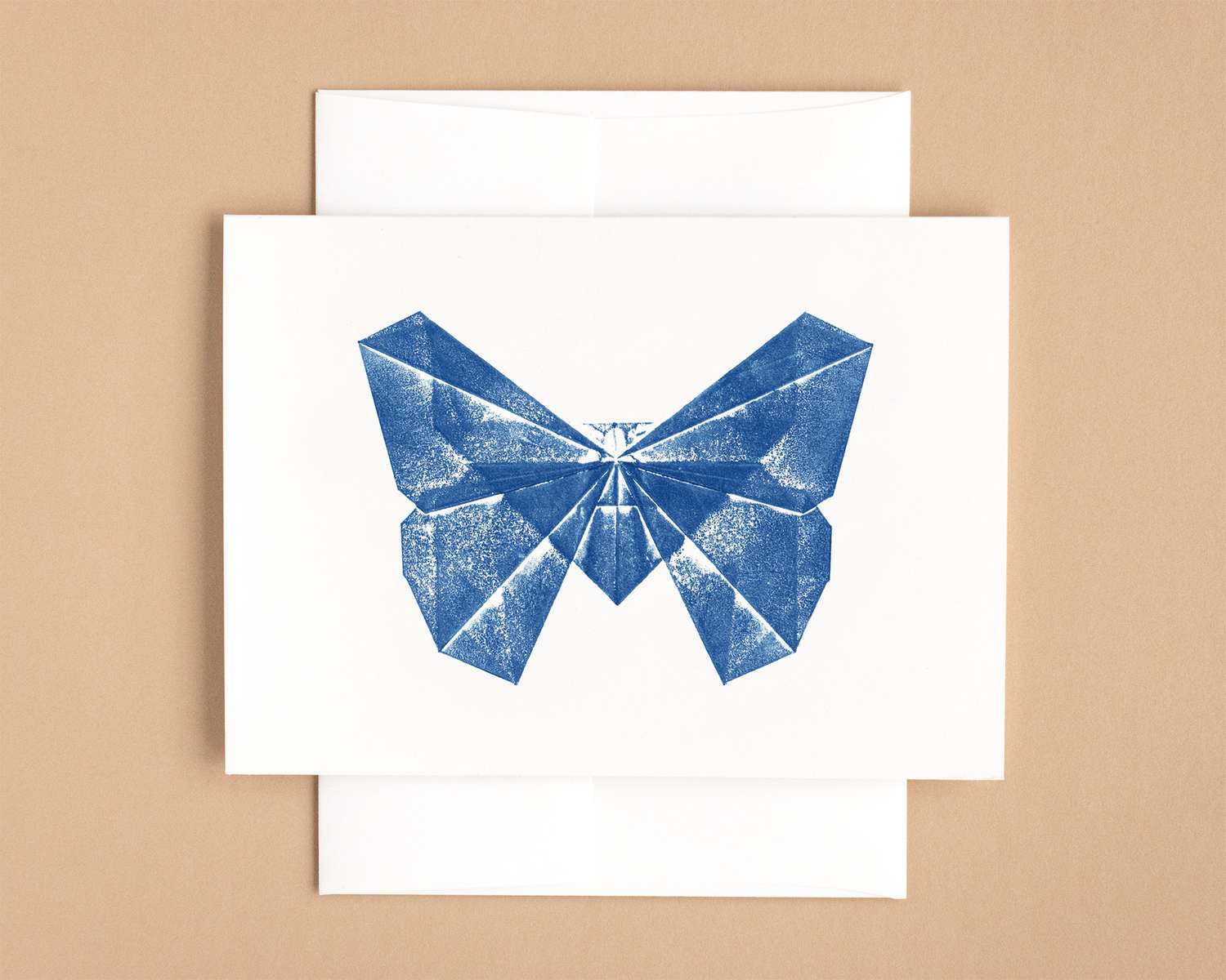 A horizontal white card depicts a bright blue butterfly, rendered in blocky and patchy triangles. The card sits on top of a white envelope, which lies on a brown backdrop.